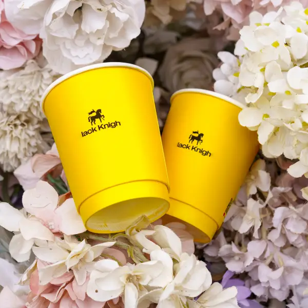 Vasos de papel amarillos impresos a medida con el logo de Black Knight colocados entre flores
